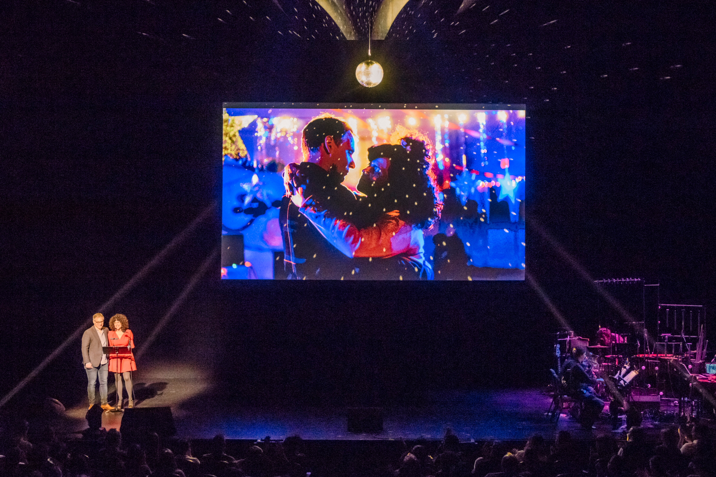 Cecilia Aldarondo and her husband perform on stage together in front of a screen with artwork of the two of them dancing.