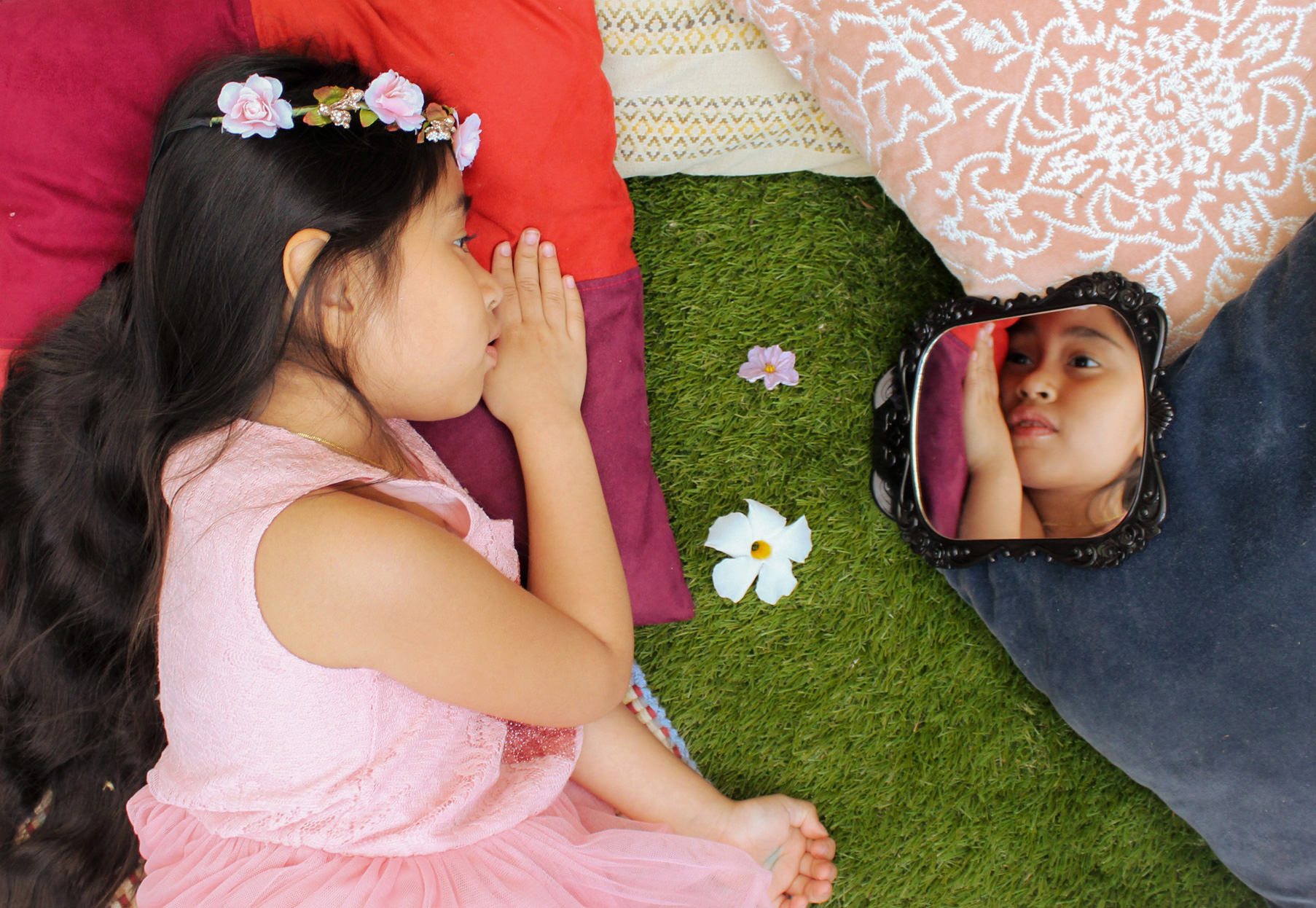 Image description: the artist’s younger sister lying on colorful pillows on grass, looking at her reflection in a small black mirror. Photo by Gaby Salazar, 18.